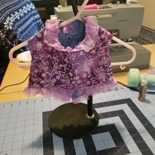 Small purple floral dress with ruffles on a miniature hanger and stand.