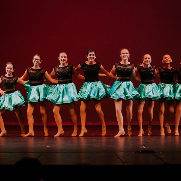 Young dancers in teal skirts and black tops performing on stage with a red backdrop.