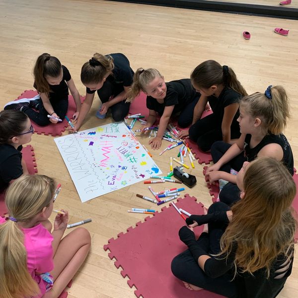 Young girls gather around a poster, drawing and coloring together on a wooden floor.