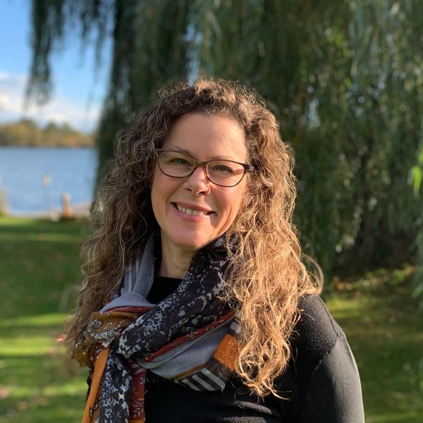 Smiling woman with curly hair and glasses outdoors near a lake and trees.