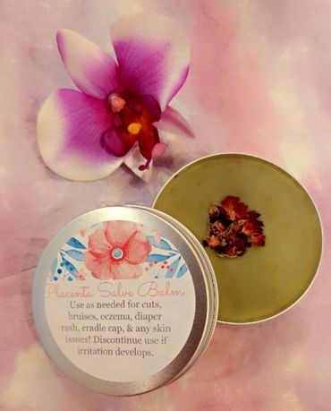 A tin of placenta salve balm with dried flowers and an orchid on a pink background.