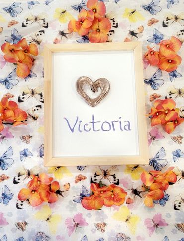 A framed artwork with a heart and the name Victoria, surrounded by orange flowers on butterfly-patterned fabric.