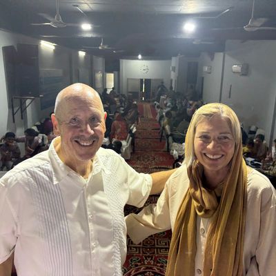 Two smiling people posing inside a room with others in the background.
