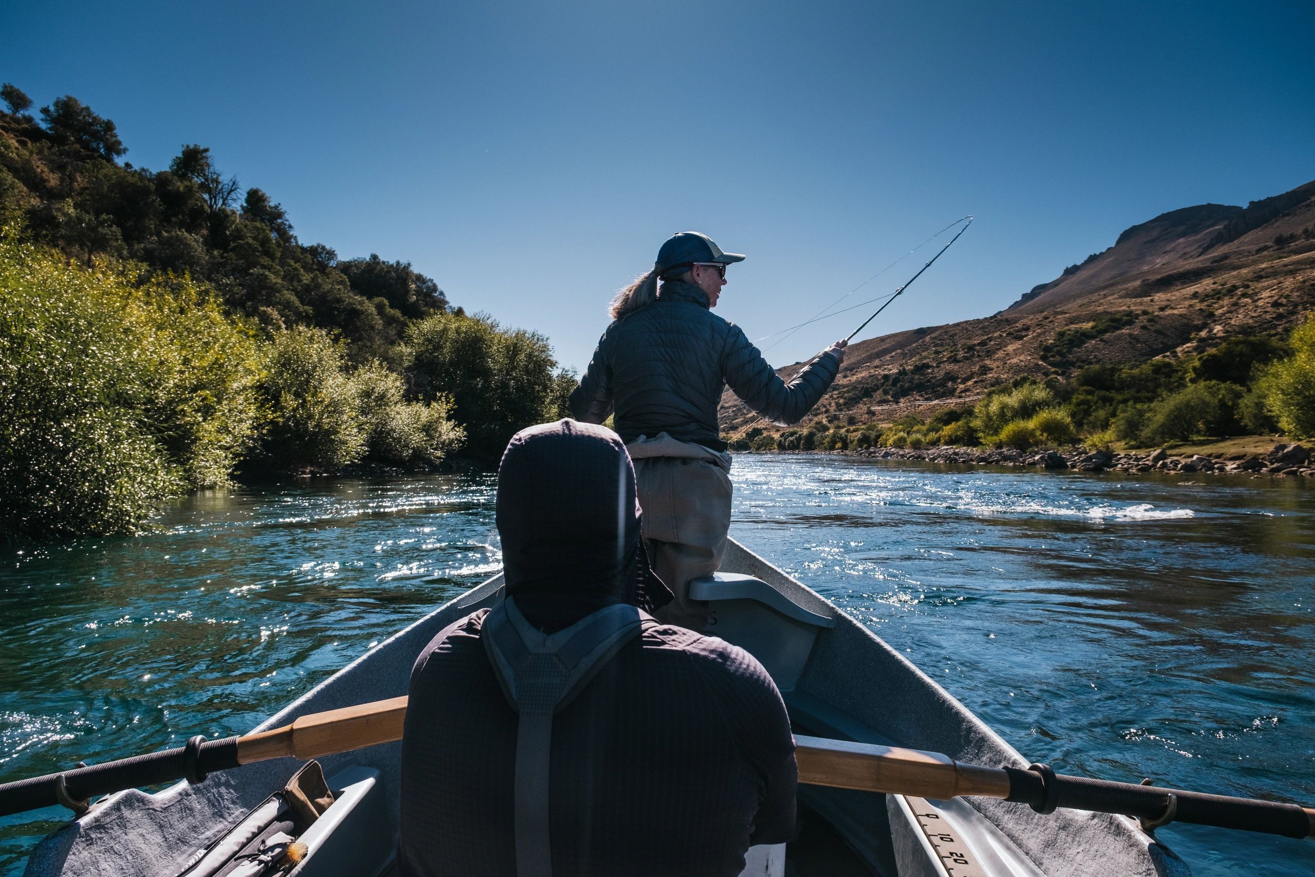 Two people fishing from a boat on a sunny river surrounded by hills and trees.