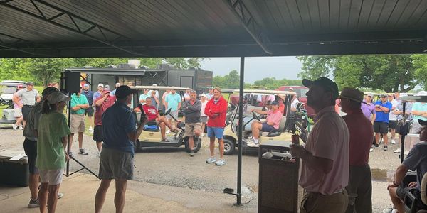 Group of people gathered under a shelter near golf carts, engaged in an outdoor event.
