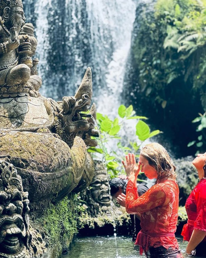 People performing a ritual near a waterfall with ornate stone statues.