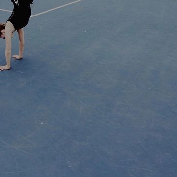 A girl in a black leotard doing a handstand on a blue tennis court.