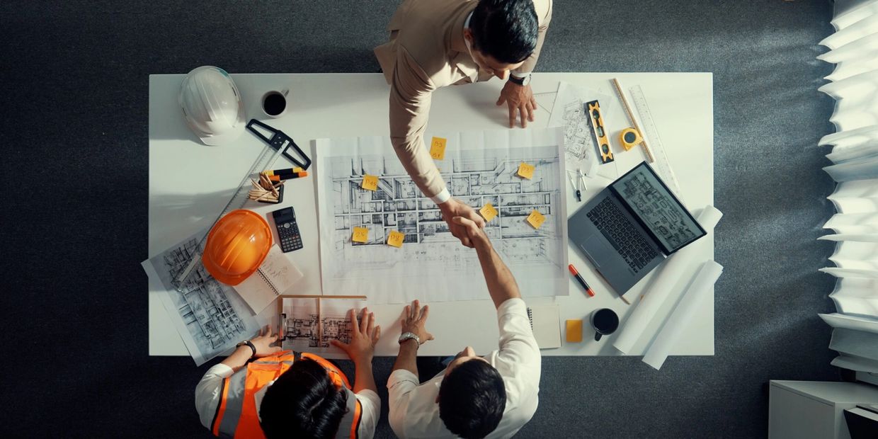 Architects and engineers shaking hands over building plans on a desk.