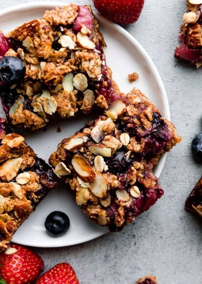 Oatmeal berry bars with nuts on a white plate surrounded by fresh strawberries and blueberries.