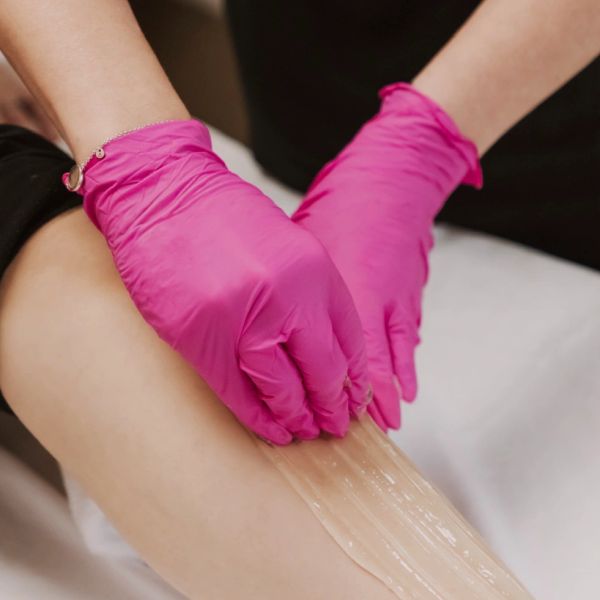 Hands in pink gloves applying wax on a leg for hair removal.