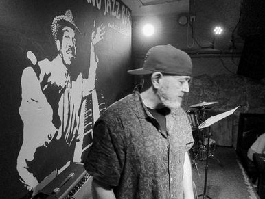 Man in a cap stands near a jazz-themed mural in a dimly lit room.