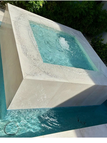 Modern square hot tub with water bubbling inside, surrounded by a pool and greenery.