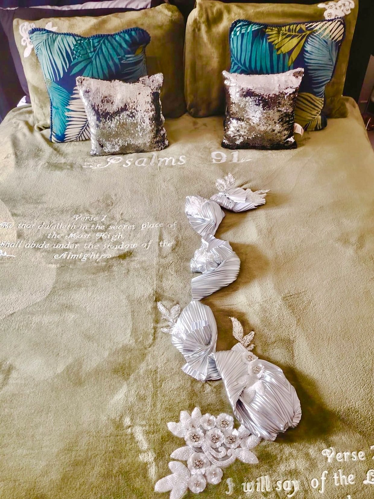 A neatly decorated bed with embroidered Psalms 91 verse and shiny cushions.