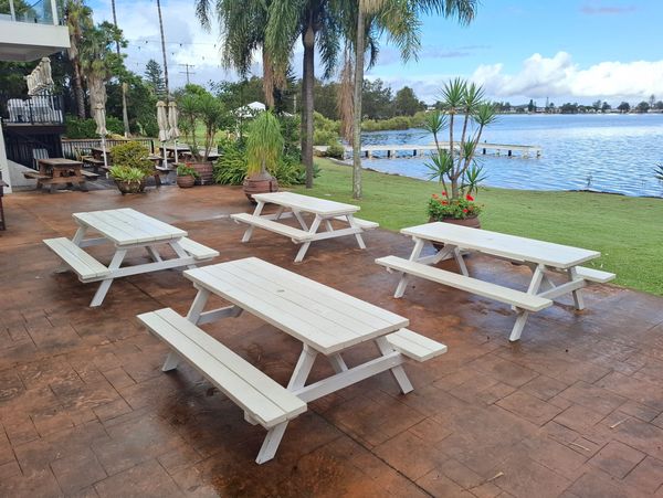 White picnic tables on a patio by a lake with palm trees and blue sky.
