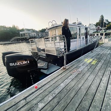 A person stands on a docked boat at sunset near a small harbor.