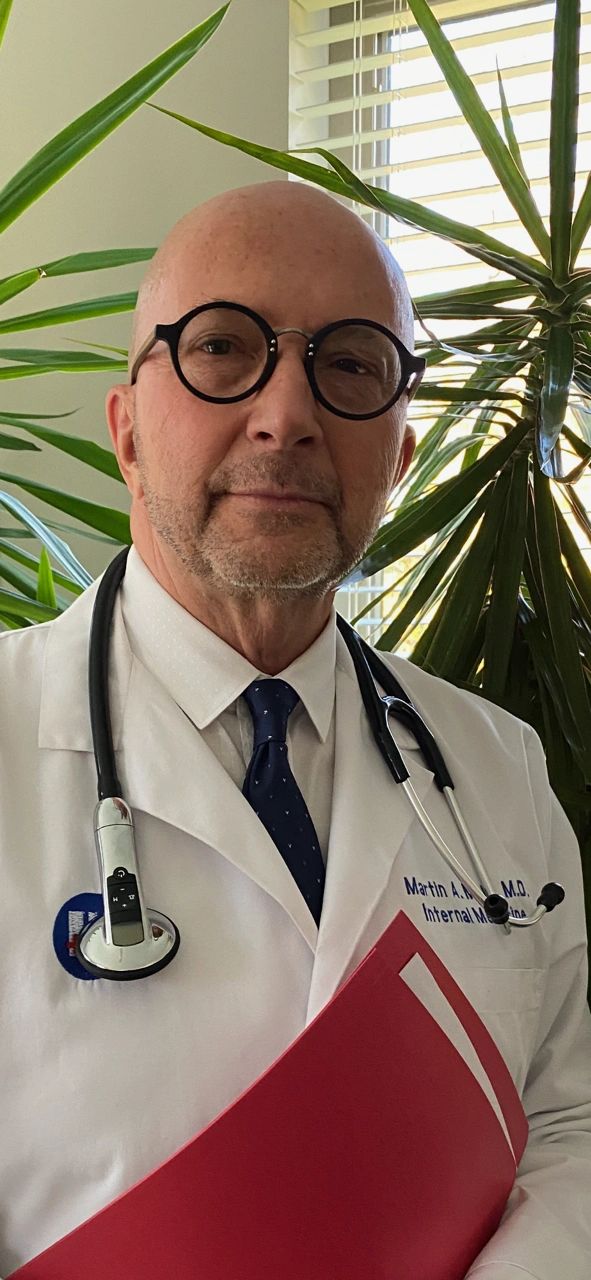 A doctor with glasses and a stethoscope stands near plants holding a red folder.