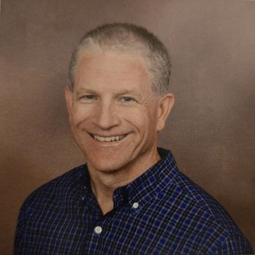 Smiling middle-aged man in a blue plaid shirt against a brown background.