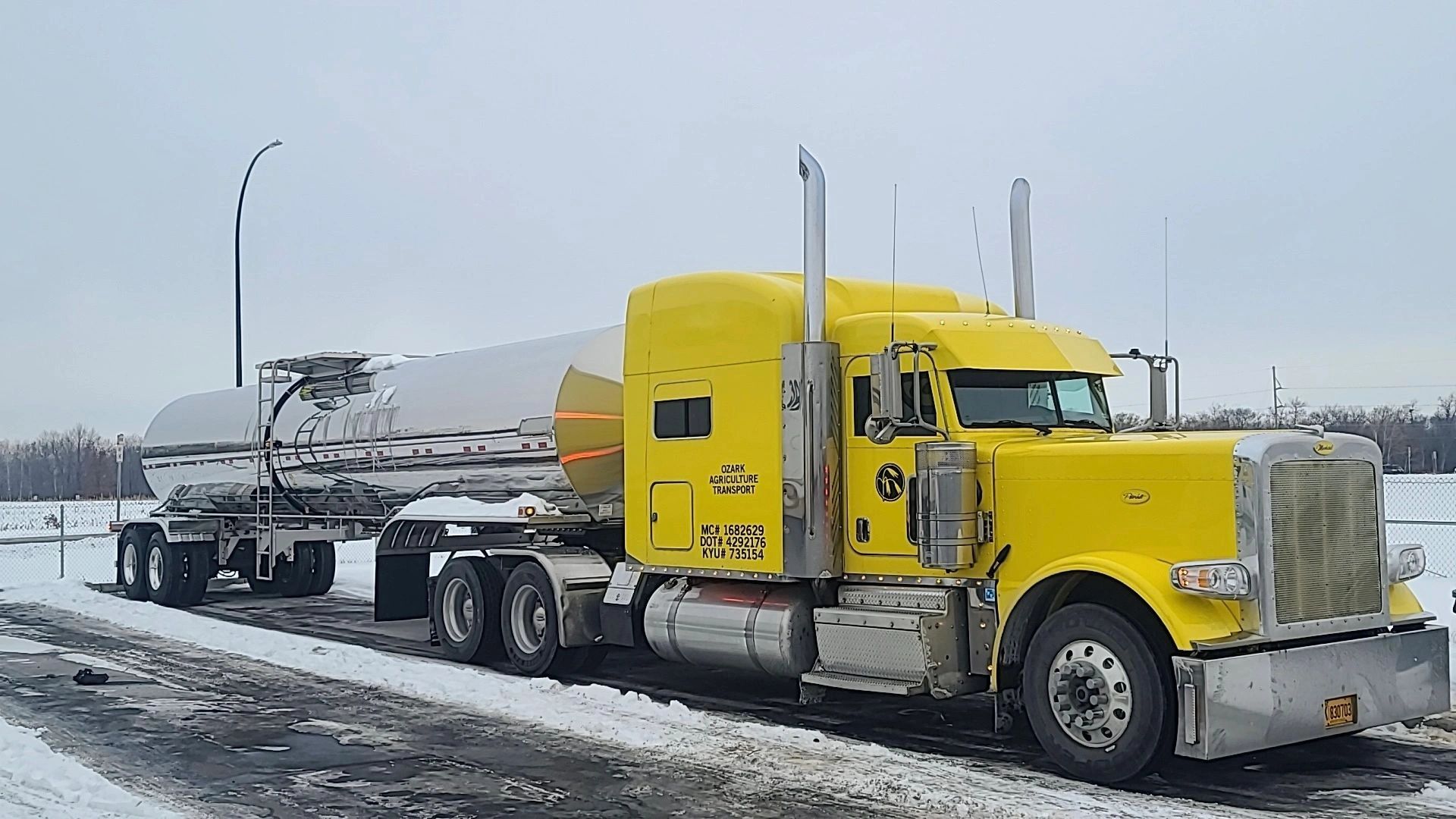 A yellow semi-truck with a shiny tanker trailer on a snowy road.