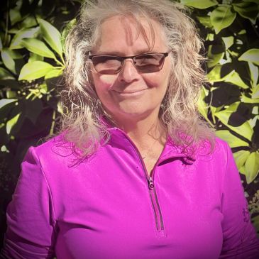 Smiling woman in a bright pink zip-up top with glasses, standing outdoors.