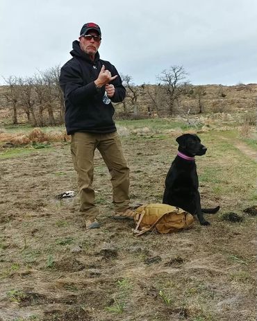 A man in outdoor gear with a black dog sitting beside a backpack in a field.