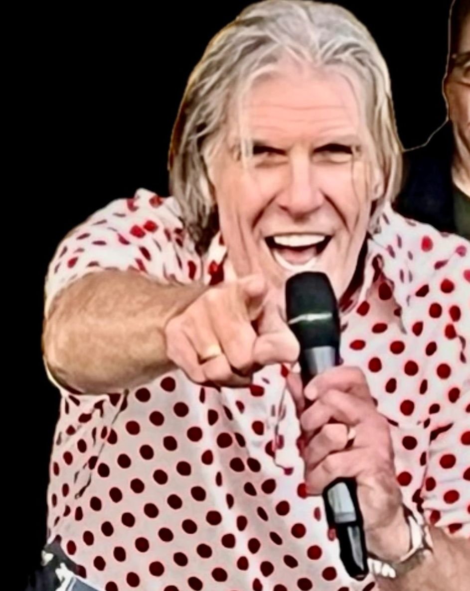 Man in a red polka dot shirt pointing and holding a microphone, smiling enthusiastically.