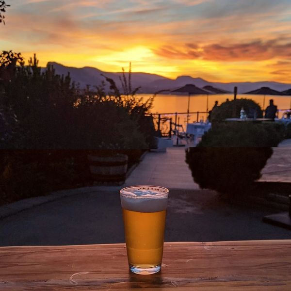 A pint of beer on a table with a vibrant sunset over mountains and water in the background.