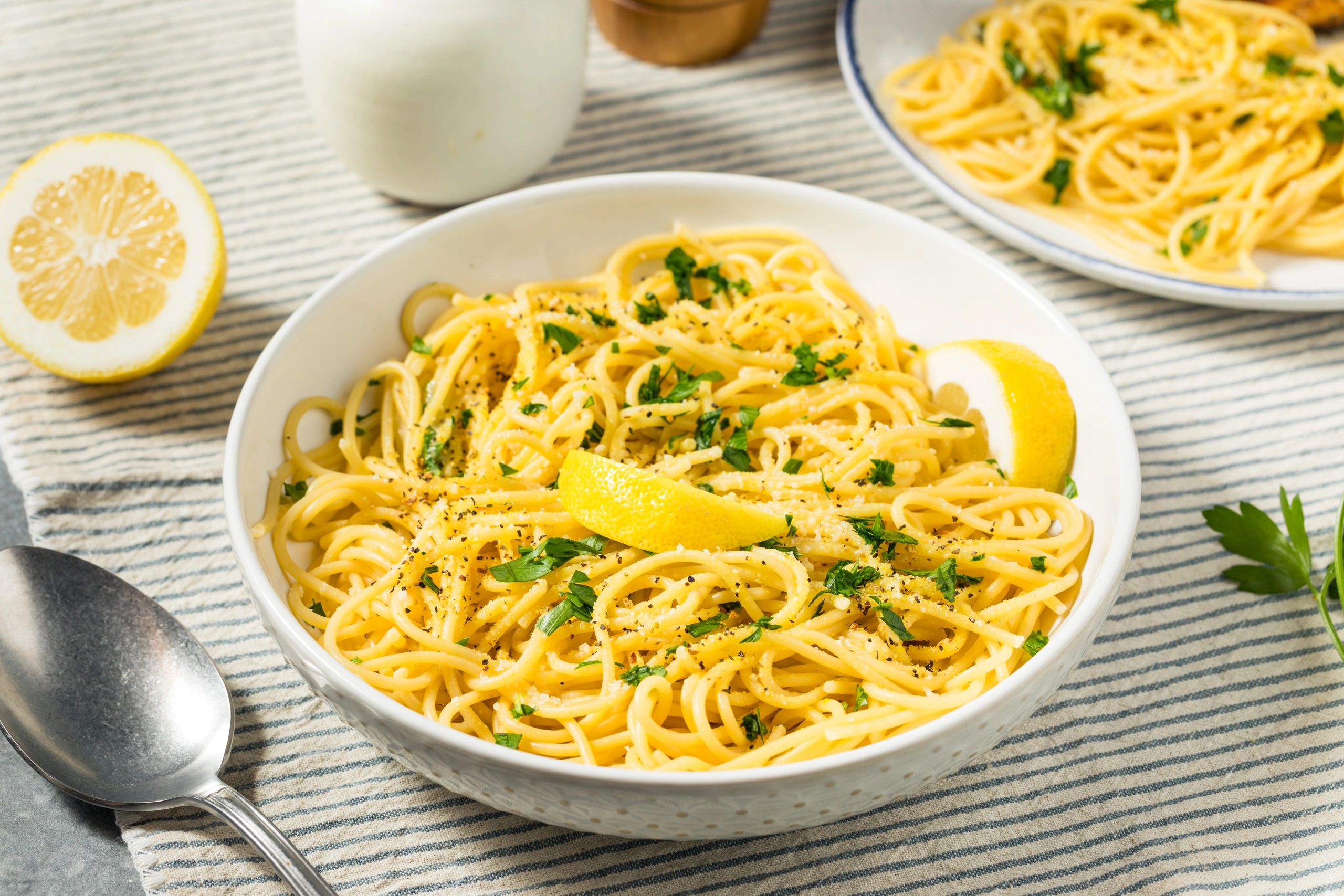 A white bowl is filled with bright yellow spaghetti, garnished with chopped parsley and black pepper