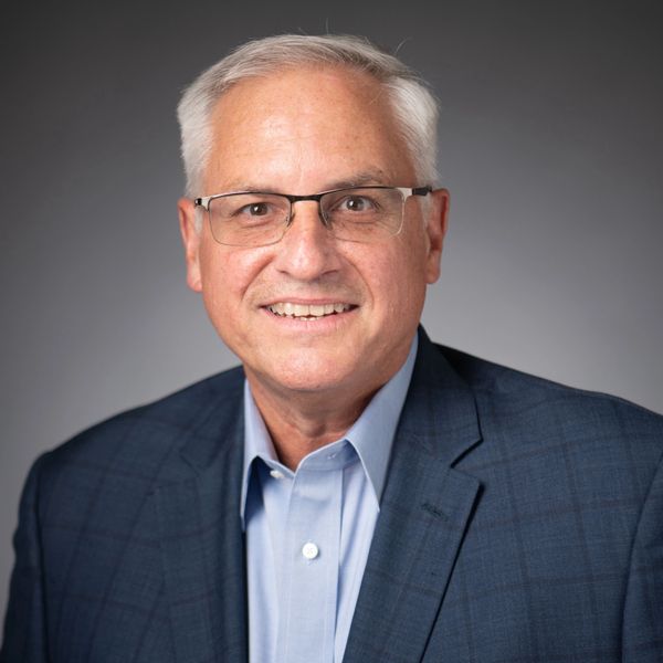 Smiling older man in glasses with gray hair wearing a blue suit jacket and light blue shirt.
