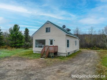 Small beige house with green roof and wooden steps, surrounded by trees and a dirt driveway.