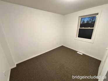 Empty room with white walls, brown carpet, and a window at dusk.