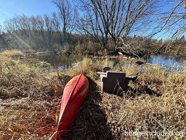 A red canoe beside a chair and table near a calm river in autumn.