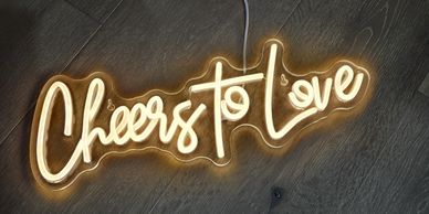 Glowing neon sign reads 'Cheers to Love' on wooden floor.