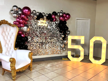 Elegant 50th birthday party setup with gold and white chair, silver backdrop, and balloon decorations.