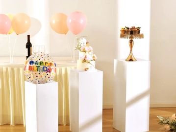 Three white pedestals displaying birthday decorations and desserts in a bright room.