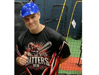 Man wearing a blue helmet and a baseball-themed shirt smiles indoors.
