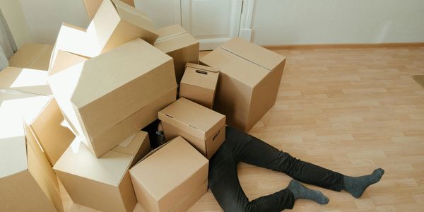 a stressed male customer laid under a heap of messy boxes on the floor in a room