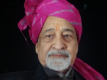 Older man wearing a pink turban and black clothing.