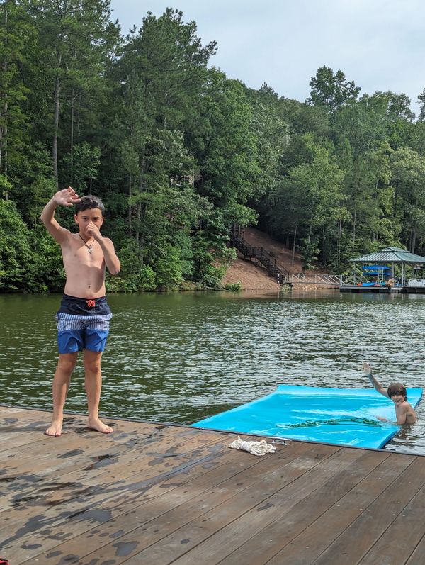 Two boys enjoying a day by the lake with one on the dock and the other in the water waving.