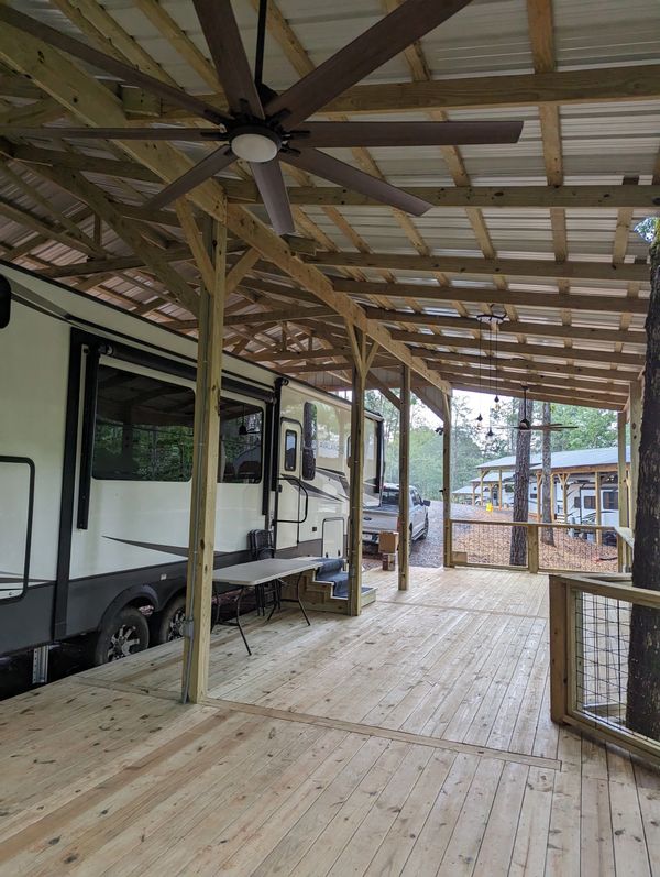 Covered wooden deck with ceiling fans next to RVs in a wooded area.