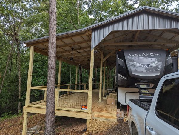 Wooden covered porch next to a parked RV and truck in a forested area.