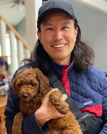 A smiling man in a cap holding a fluffy brown puppy indoors.