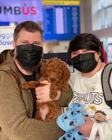 Couple wearing black masks holding a brown puppy indoors.