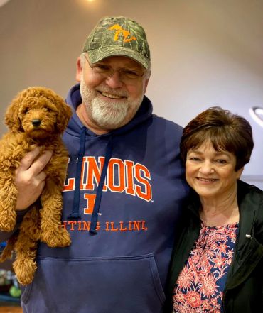 Smiling couple holding a fluffy brown puppy indoors.