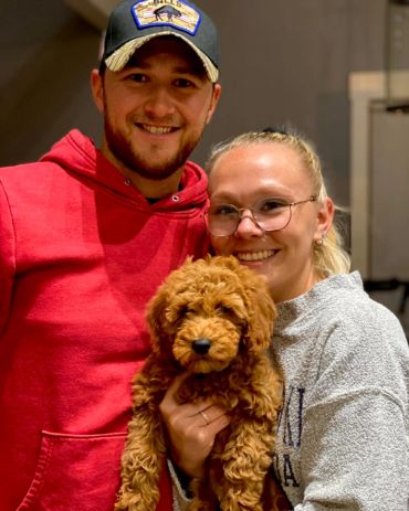 Smiling couple holding an adorable curly-haired puppy indoors.