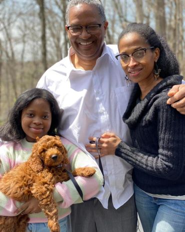 A smiling family of three with a fluffy brown dog outdoors.