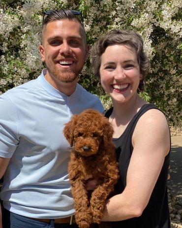 Happy couple holding a brown fluffy puppy outdoors in front of white flowers.