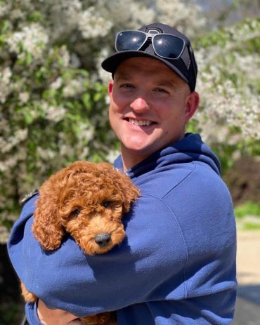 Man in blue hoodie holding a curly brown puppy outdoors.