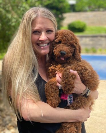 Woman smiling and holding a fluffy brown puppy outdoors.