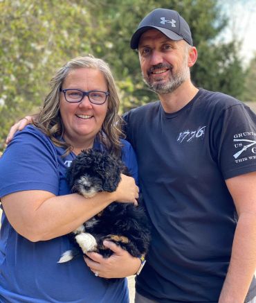 Smiling couple holding a fluffy black and white puppy outdoors.