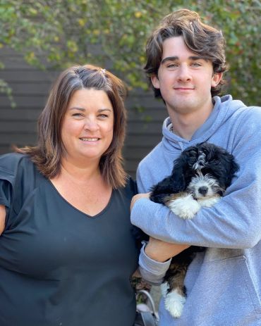 A woman and a young man smiling, the man holding a fluffy black and white puppy.